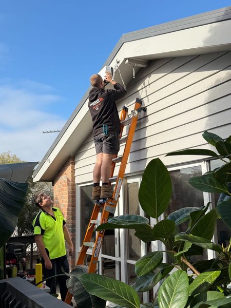 A powerband electrical installer on a ladder working on the eaves of wayne's home, a colleague spotting below