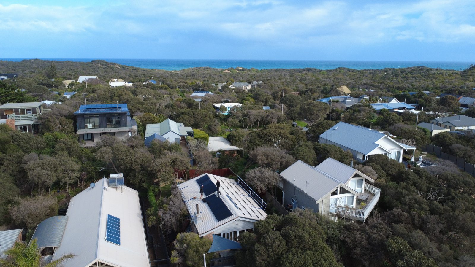 Wide aerial view of blairgowrie homes along the mornington peninsula coastline, ocean in the distance, installers on a roof mid-install
